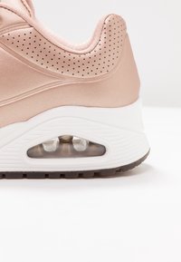 Ljusrosa träningsskor med slät tyg, perforerade detaljer, en vit mellansula och en synlig Air-dämpningsenhet i hälen.