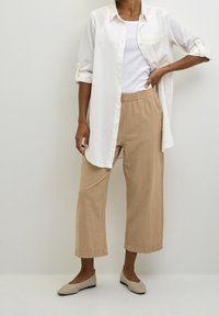 Chemise boutonnée en lin blanc portée sur un débardeur blanc, associée à un pantalon beige court et des chaussures légères à enfiler de couleur claire. Textures lisses, coupe décontractée.