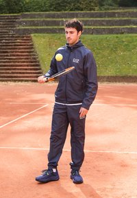 Fato de treino atlético azul com logótipo, sapatos pretos, segurando uma raquete de ténis. Bola de ténis amarela no ar em um campo de terra batida com fundo verde.