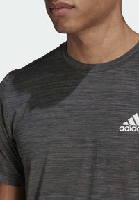 Grå atletisk t-shirt med rund halsringning, tillverkad av lätt och texturerad tyg. Har en vit Adidas-logotyp på vänster bröst.