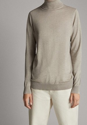 Pull à col roulé beige à manches longues porté rentré dans un pantalon blanc cassé, visible du cou jusqu'aux hanches sur un fond gris uni.