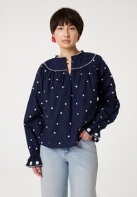 Navyblaue Bluse mit weißer Blumen- und Herzstickerei, einem runden Kragen, Knopfverschluss und weit ausgestellten Ärmeln mit Verzierungen. Aus weichem, leichtem Material.