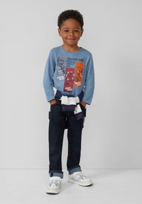 Blauwe langermige shirt met grafische ontwerpen, donkere denim jeans, en witte sneakers met beige accenten. Trui om de taille geknoopt.