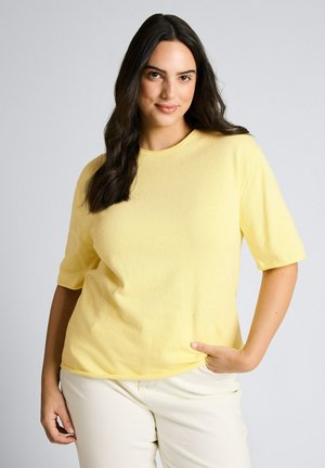 Frau mit langen dunklen Haaren trägt ein gelbes kurzärmeliges Oberteil und helle Hosen, steht mit einer Hand in der Tasche.