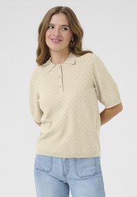 Beige polo triko s krátkým rukávem, s texturovaným diamantovým vzorem a třemi knoflíky u límce. V kombinaci se světle modrými džíny.