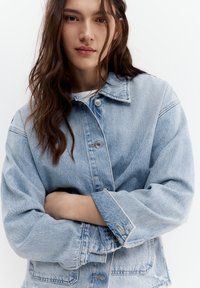 Junge Frau mit langen braunen Haaren, trägt eine hellblaue übergroße Jeansjacke, Arme verschränkt, neutraler Gesichtsausdruck, weißer Hintergrund.