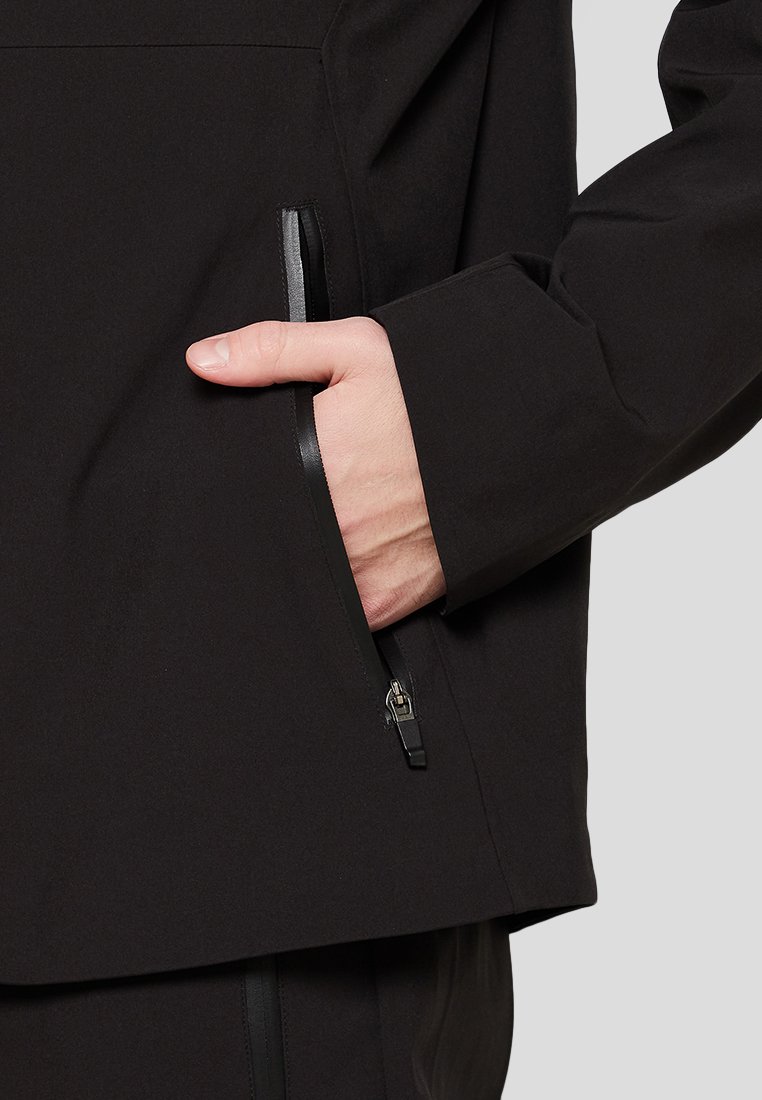 Giacca nera con tasca anteriore con zip, tessuto testurizzato, una leggera lucentezza e una vestibilità rilassata, con una mano infilata in una tasca laterale.