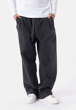 Persona che indossa jeans neri larghi con coulisse in vita, sneakers bianchi e una camicia bianca a maniche lunghe, mani nelle tasche, in piedi su sfondo bianco.