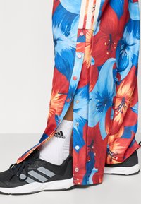 Φλοράλ αθλητικό παντελόνι με σχέδια από μπλε και κόκκινα λουλούδια, με κλείσιμο με κουμπιά τύπου snap και τρεις κάθετες ρίγες. Μαύρα αθλητικά παπούτσια με γκρι λεπτομέρειες.