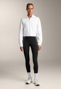 Felpa bianca corta con zip e colletto alto, abbinata a leggings neri con cuciture laterali bianche e scarpe sportive grigie con dettagli bianchi.