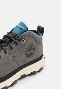 La bota de senderismo de ante gris presenta un cuello acolchado azul, una suela de goma negra y un logotipo grabado en el lateral. También incluye una malla texturizada y cordones.