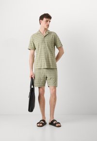 Green striped short-sleeve shirt and matching shorts made from textured fabric. The model holds a black bag and wears slide sandals.