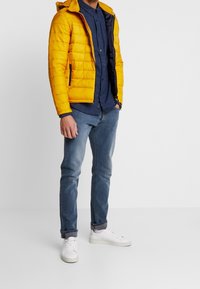 Gul puffjakke med lynlås, marineblå inderfor, båret over en marineblå ternet skjorte, parret med lyseblå jeans og hvide sneakers.