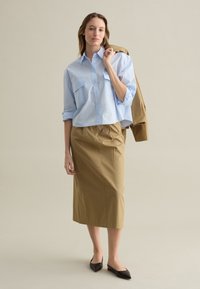 Camisa de botones azul claro con dos bolsillos frontales, combinada con una falda beige de longitud a la rodilla y bailarinas negras puntiagudas, sosteniendo una chaqueta color marrón claro.