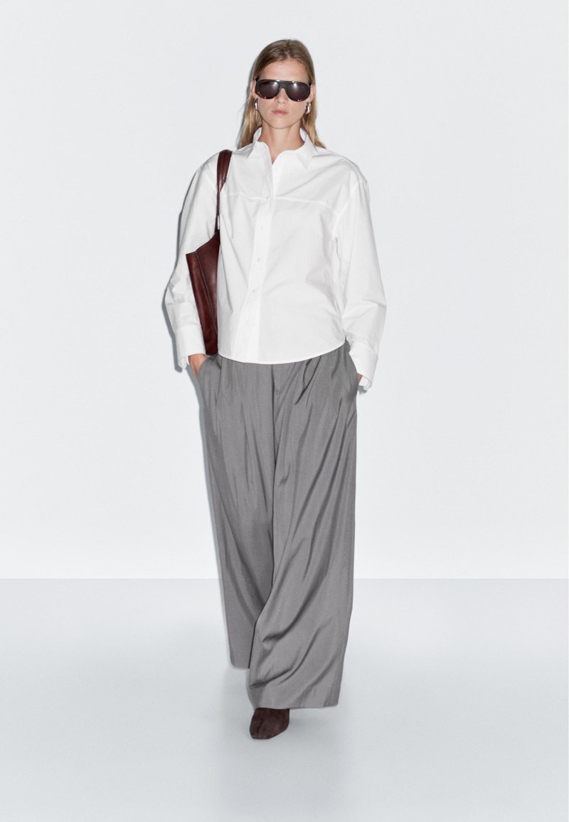 Donna che cammina avanti indossando una camicia bianca oversize con bottoni, pantaloni larghi grigi, occhiali da sole scuri, portando una borsa a tracolla marrone, con le mani nelle tasche.