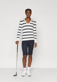 Suéter branco com riscas horizontais azuis-marinho, decote em V, textura canelada, combinado com calções azuis-marinho até ao joelho e ténis brancos. Taco de golfe na mão.