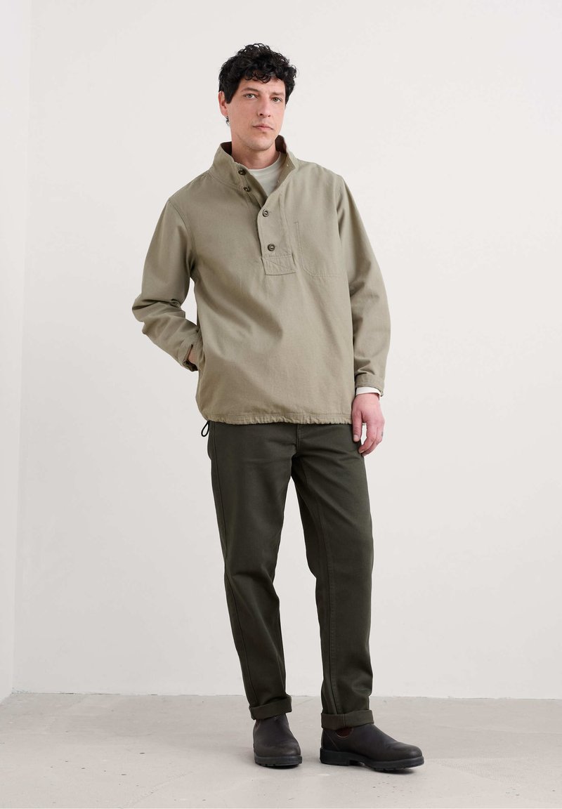Suéter de manga larga en verde oliva claro con una abertura de medio botón y bolsillo frontal, combinado con pantalones verde oscuro y calzado oscuro.