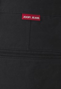 Jeans in denim nero con un'etichetta di tessuto rossa che riporta la scritta "JOOP! JEANS", caratterizzati da una superficie liscia e un classico design a cinque tasche.