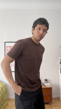 Homme aux cheveux courts et foncés, portant un t-shirt marron et un pantalon noir, debout dans une pièce avec des meubles en bois et des œuvres encadrées au mur.