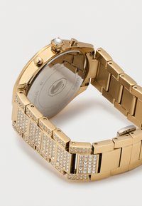 Ρολόι χειρός σε χρυσό χρώμα με μπρασελέ διακοσμημένο με κρύσταλλα και τρία πλευρικά κουμπιά, σε θέα από πίσω σε λευκό φόντο.