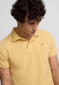 Camisa polo de algodón amarilla con cuello clásico, mangas cortas y un pequeño logotipo negro en el pecho. Textura suave y diseño ajustado.