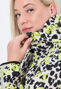 Donna bionda che sistema il cordino di una giacca con colletto alto, decorata con un motivo leopardato verde, nero e bianco.