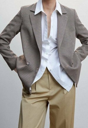 Personne portant un blazer marron à motif pied-de-poule sur une chemise blanche boutonnée et un pantalon plissé beige, les mains dans les poches.