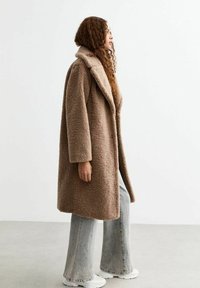 Brun teddyjacka med en plyschig, texturerad yta, med en stor krage, öppen framsida och långa ärmar, ihop med urvuxna utsvängda jeans och vita skor.