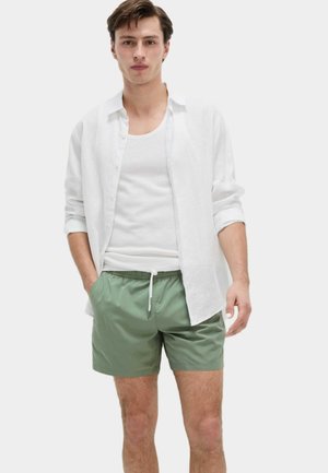 Modelo vistiendo una camisa de lino blanca sobre una camiseta sin mangas blanca y pantalones cortos de baño verdes. Los pantalones cortos cuentan con una cintura elástica y cordones.
