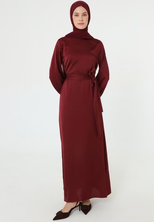 Donna in piedi con le mani dietro la schiena, indossa un abito lungo bordeaux a maniche lunghe, hijab abbinato e tacchi neri a punta.