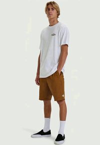 White short-sleeve t-shirt with black logo, brown shorts with pockets, black slip-on shoes, and white crew socks. Casual fit, smooth texture.