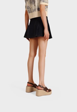 Donna che indossa una mini gonna plissè nera, top beige con motivo, e sandali zeppa marroni con cinturini alla caviglia, in piedi contro uno sfondo semplice.