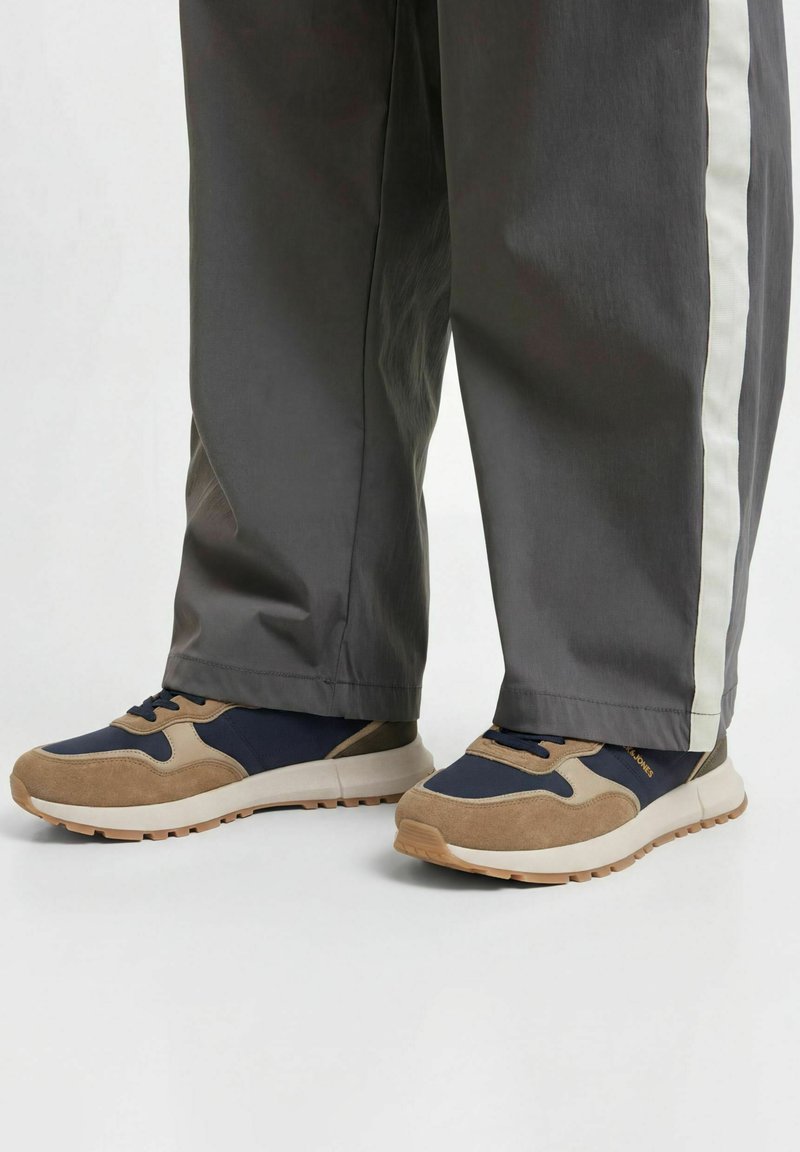 Personne portant un pantalon gris avec des bandes blanches sur les côtés et des baskets marron et bleu marine debout sur un sol blanc.