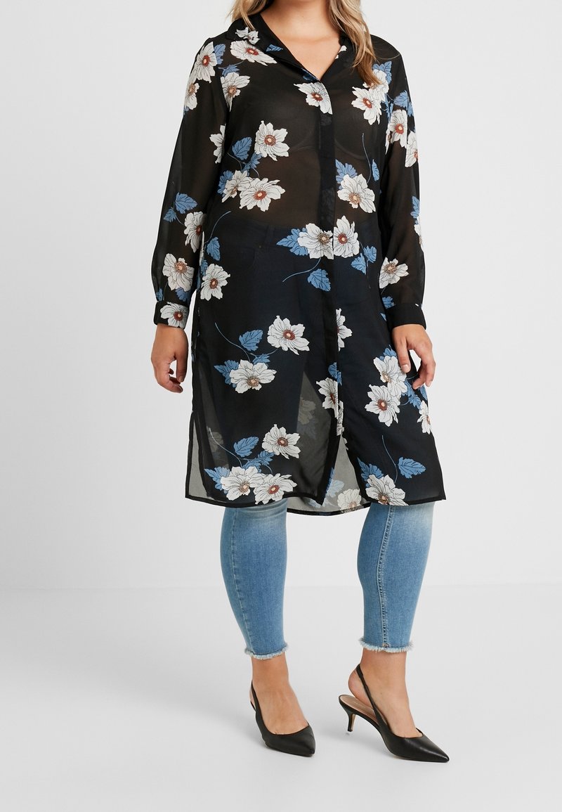 Sort gennemsigtig tunika med blomsterprint, der har hvide og blå blomster, parret med blå skinny jeans og sorte spidse hæle.
