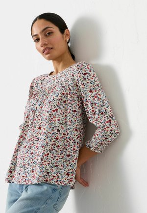 Woman with dark hair tied back wears floral long-sleeve blouse and light blue jeans, leaning against a white textured wall.