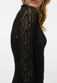 Vestido negro de encaje con mangas largas, que presenta un patrón floral texturizado. La tela es semitransparente, revelando sutilmente la piel debajo.