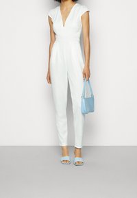 Mono blanco con mangas cortas, escote en V profundo y cintura ajustada; combinado con un bolso de mano azul claro y sandalias deslizantes a juego.