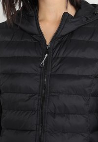 Chaqueta acolchada negra con capucha, cierre frontal y tejido brillante. Las líneas de costura crean un diseño estructurado con una textura suave.