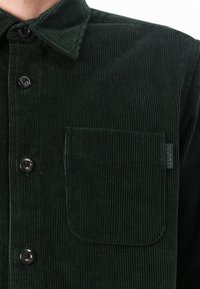 Dark green corduroy shirt with a button-up front, textured fabric, and a chest pocket, featuring a small branded label on the pocket.