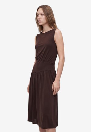 Donna con capelli castano chiaro che indossa un vestito senza maniche marrone scuro fino al ginocchio, in piedi davanti a uno sfondo chiaro e uniforme.
