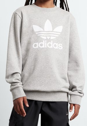 Person trägt ein hellgraues Adidas-Sweatshirt mit großem weißem Logo und schwarze Hose, steht vor einem einfarbigen Hintergrund.