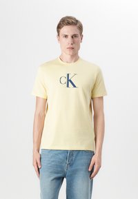 Žuta pamučna majica s kratkim rukavima i okruglim izrezom, s mornarsko plavim CK logotipom. Uparena s svijetloplavim trapericama.