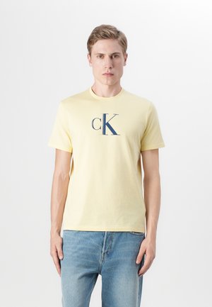 Camiseta de algodón amarilla con un logo cK en azul marino, de manga corta y cuello redondo. Combinada con jeans de mezclilla azul claro.