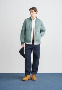 Chaqueta acolchada color verde azulado con bolsillos laterales, llevada sobre una camisa blanca. Pantalones azul oscuro y botas robustas color tostado completan el conjunto. Gorra negra en la mano.