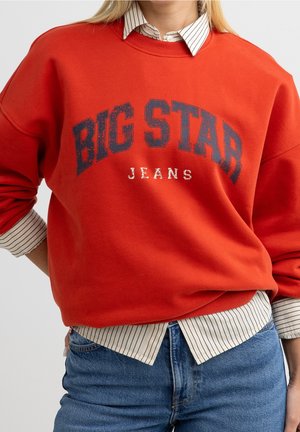 Osoba nosząca czerwoną bluzę "BIG STAR JEANS" na białej koszuli w cienkie paski i niebieskich dżinsach, ręka na biodrze, na jednolitym tle.
