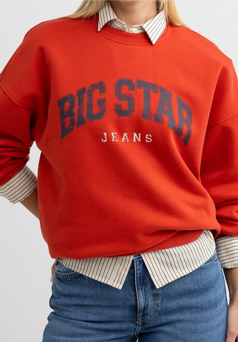 Personne portant un sweat-shirt rouge "BIG STAR JEANS" par-dessus une chemise blanche à fines rayures et un jean bleu, main sur la hanche, sur un fond uni.