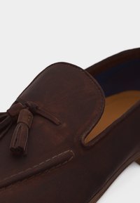 Mocassin en cuir marron foncé avec des pampilles et des coutures détaillées sur la partie supérieure, vue partielle de côté et de dessus.