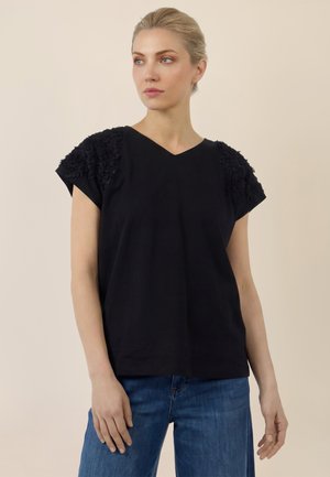 Blonde Frau, die ein schwarzes Top mit kurzen Ärmeln und strukturierten Schulterdetails sowie blaue Jeans trägt, leicht nach rechts blickend.