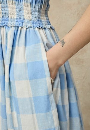 Main dans la poche d'une robe à carreaux bleus et blancs avec une taille smockée, révélant un tatouage de papillon sur l'avant-bras et un bracelet de perles au poignet.