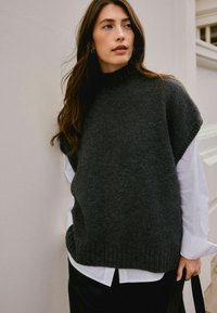 Woman with long brown hair wearing a dark gray sleeveless sweater over a white shirt, holding a black bag, standing against a light wall.